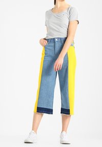 Denim culottes featuring a light blue wash with bright yellow side panels, raw hem, and a high-rise fit, paired with a gray t-shirt.