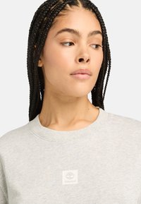 Svetle šedá mikina s kulatým výstřihem, která má uprostřed malé čtvercové logo Timberland. Hladká textura a ležérní design.