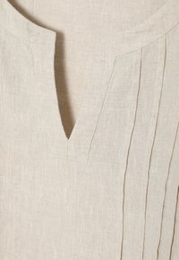 Beige lněná košile se dvěma výstřihem do V a jemným vertikálním skladem na jedné straně. Texturovaná látka s měkkým, přirozeným vzhledem.