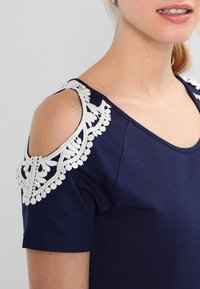 Camiseta azul marino con diseño de hombros descubiertos, presenta detalles de encaje blanco en los hombros. Tela suave con una textura lisa.