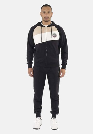 Uomo che indossa pantaloni della tuta neri e una felpa con cappuccio con zip nera, beige e bianca, con sneaker bianche, in piedi davanti a uno sfondo chiaro uniforme.