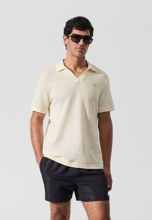 Krém színű, texturált kötött anyagból készült galléros póló rövid ujjakkal és V-nyakú gallérral. Fekete rövidnadrággal és sötét napszemüveggel párosítva.