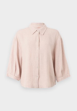 Jeune femme debout pieds nus, vêtue d'une chemise ample rose à carreaux boutonnée et d'un short assorti, sur un fond blanc uni.