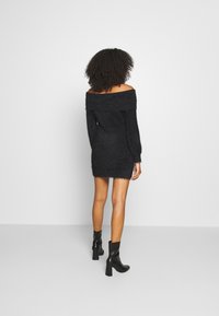 Robe noire en maille épaules dénudées avec manches longues et ourlet effiloché, portée avec des bottines noires à talons, sur un fond uni.