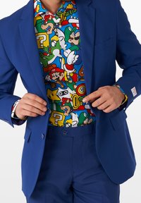 Blazer blu sopra una camicia multicolore con bottoni, caratterizzata da disegni grafici di personaggi dei videogiochi, tra cui Mario e Yoshi.