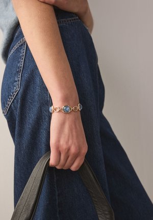 Gold bracelet featuring blue gemstones and oval links, worn on a wrist. Hand holds a black leather bag, with denim fabric visible.