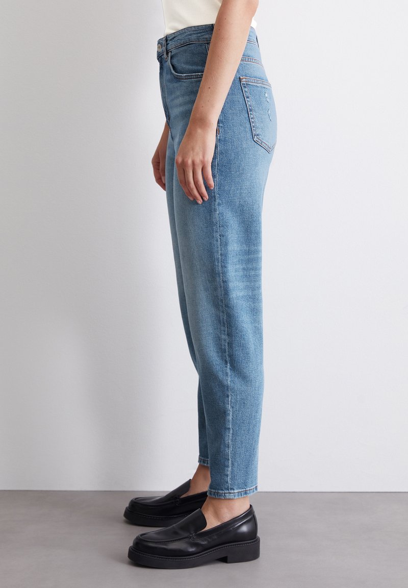 Hochgeschnittene hellblaue Jeans aus Denim mit geradem Bein. Verfügt über ein Fünf-Taschen-Design und minimale Abnutzungsspuren. Getragen mit schwarzen Slippern.