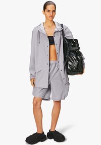 Jasnoszara kurtka przeciwdeszczowa z kapturem noszona na czarnym topie crop, w zestawieniu z jasnoszarymi szortami, czarną torbą oversized oraz fakturowanymi czarnymi butami.