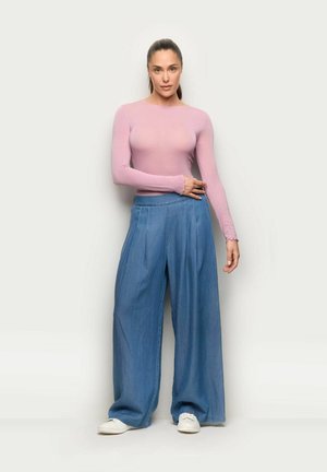 Femme debout contre un fond uni, portant un haut rose ajusté à manches longues, un jean bleu à jambes larges et des baskets blanches.