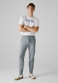 Biele grafické tričko s nápisom "RED BULL RACING", v kombinácii so sivými teplákmi a pieskovými športovými topánkami s čiernymi akcentmi.