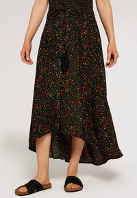 Jupe maxi noire à motifs floraux rouges et jaunes, taille smockée, ceinture ajustable et ourlet asymétrique. Portée avec des sandales à glissière noires.