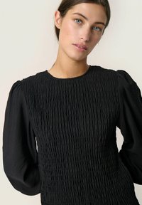Black dress with textured, smocked bodice and puffed sleeves. Smooth fabric contrasts with the gathered texture of the top.