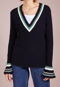 Námořnický pletený svetr s výstřihem do V, s zelenými a bílými pruhy na límci a rozšířenými lemy rukávů, doplněný modrými džínami.