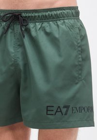 Tumši zaļi peldšorti ar elastīgu jostasvietu un melnu auklu. Rāda gludu tekstūru un apdruku ar "EA7 EMPORIO" logotipu.