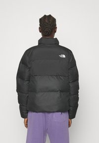 Czarna kurtka puchowa z wysokim kołnierzem, poziomymi przeszyciami i małym białym logo na górnej lewej części pleców. Miękkie, matowe wykończenie.