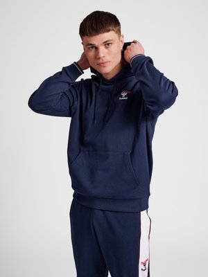 Jonge man past de capuchon van een donkerblauwe hoodie met hummel-logo aan, draagt bijpassende donkerblauwe joggingbroek met witte zijstrepen en logo's.