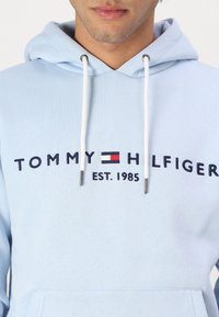 Felpa leggera azzurra con tasca frontale, cordini bianchi e logo del marchio ricamato in blu navy con accenti rossi e bianchi.