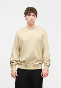 Μπεζ φούτερ με στρογγυλή λαιμόκοψη και ριμπ μανσέτες. Έχει μικρό λευκό λογότυπο στο στήθος και άνετη εφαρμογή, συνδυασμένο με μαύρο παντελόνι.