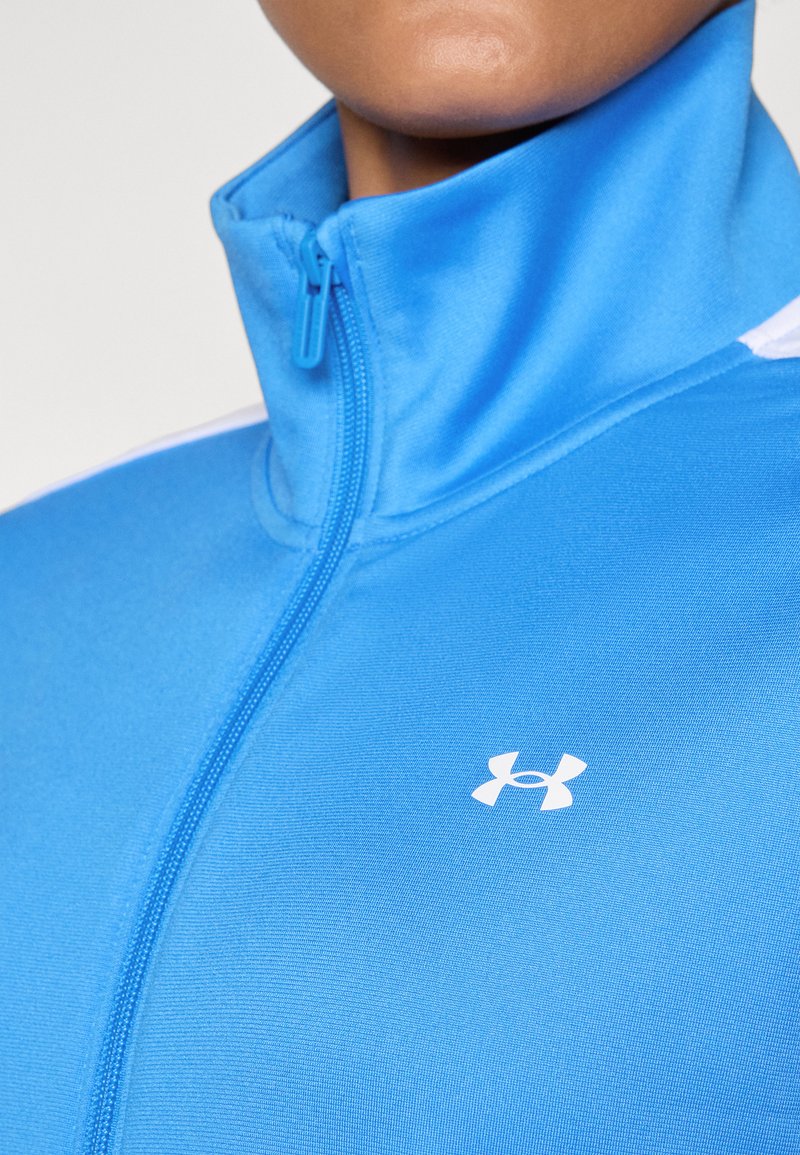 Chaqueta azul con cremallera y cuello alto, hecha de una tela suave que presenta un logo blanco en el pecho y acentos blancos en contraste.