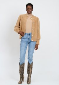 Cardigan beige soffice con bottoni in perla rotondi, indossato sopra un dolcevita chiaro, abbinato a jeans blu chiaro e stivali alti con motivo.