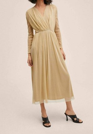 Largo vestido beige con un frontal envolvente y mangas largas, que presenta una cintura fruncida y una falda suave y ligera. Combinado con sandalias de tacón negras.