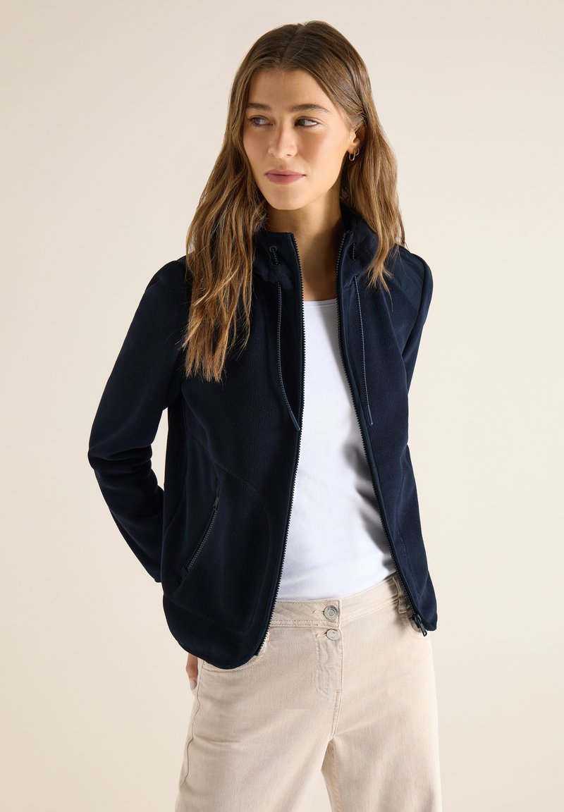 Femme aux longs cheveux bruns portant une veste zippée bleu marine, une chemise blanche et un pantalon beige, debout avec les mains derrière le dos, regardant vers la droite.