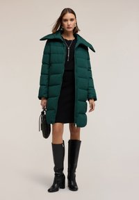 Zelená péřová bunda s velkým límcem, zipovým zapínáním a prošívanou texturou. Spárována s černými šaty a černými koženými kozačkami s krokodýlím vzorem až do kolen.