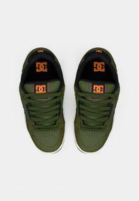 Green sneakers with a textured finish, featuring black accents, flat laces, and an orange logo on the tongue. Round toe and cushioned sole.