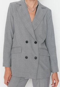 Šedý kostkovaný dvouřadový blazer s klopami a dvěma předními kapsami. Obsahuje černé knoflíky a strukturovaný střih. Doplněná texturou látky.