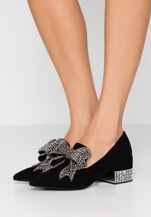 Czarne zamszowe buty na wysokim obcasie z ostro zakończonymi noskami, ozdobione dużą kokardą wysadzaną cyrkoniami oraz masywnym, dekoracyjnym srebrnym obcasem.