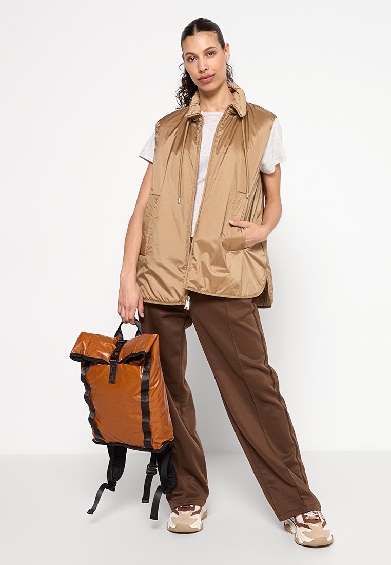 Gilet imbottito senza maniche color sabbia con due tasche frontali e colletto alto. Il modello tiene uno zaino pieghevole marrone con spallacci e dettagli neri.