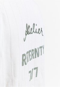 Tissu blanc avec couture verticale et texte brodé en vert indiquant « Atelier », « RTERNITY » et « 7/7 » dans différentes polices et tailles.