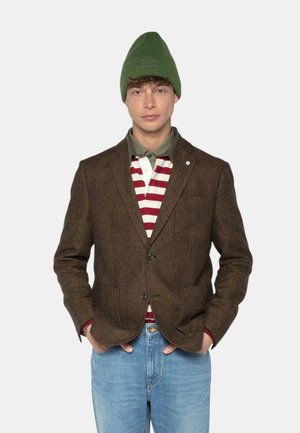 Homme portant un bonnet en tricot vert, un blazer marron, un polo rayé rouge et blanc, une chemise verte en dessous, et un jean bleu clair, les mains dans les poches.
