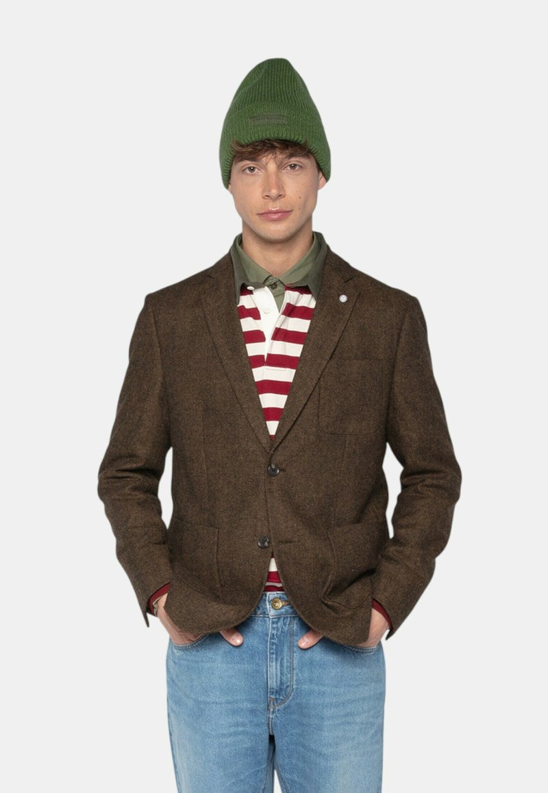 Homme portant un bonnet en tricot vert, un blazer marron, un polo rayé rouge et blanc, une chemise verte en dessous, et un jean bleu clair, les mains dans les poches.