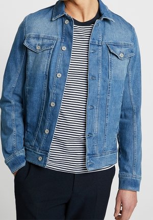 Mężczyzna ubrany w niebieską dżinsową kurtkę na czarno-białej, paskowanej koszulce i czarnych spodniach, stojący z jedną ręką w kieszeni.