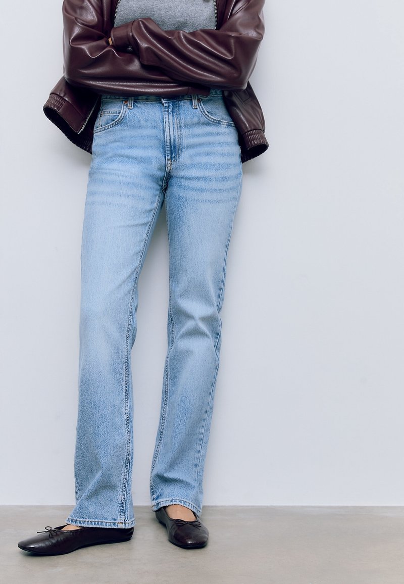 Personne portant un jean bleu clair, des ballerines noires, une chemise grise et une veste en cuir marron, debout les bras croisés contre un mur blanc.