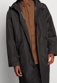 Abrigo largo negro tipo parka con capucha, que cuenta con bolsillos laterales con cremallera, una textura impermeable y un corte relajado sobre una camisa de botones marrón.