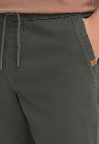 Pantalon vert foncé avec une texture douce, taille élastique et cordon de serrage. Comprend des poches latérales et une petite étiquette logo marron sur la ceinture.