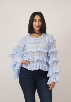 Vrouw met schouderlang donker haar, gekleed in een doorschijnende lichtblauwe blouse met ruches en donkere spijkerbroek, staand met één hand op haar heup.