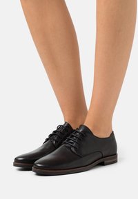 Chaussures en cuir noir à lacets au design épuré, à bout arrondi et semelle brune à profil bas, mettant en valeur une texture lisse et des coutures discrètes.