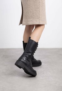 Botas de cuero negro con cordones y suela de goma gruesa, textura suave y tiradores laterales, combinadas con una falda de color marrón claro hasta la rodilla.