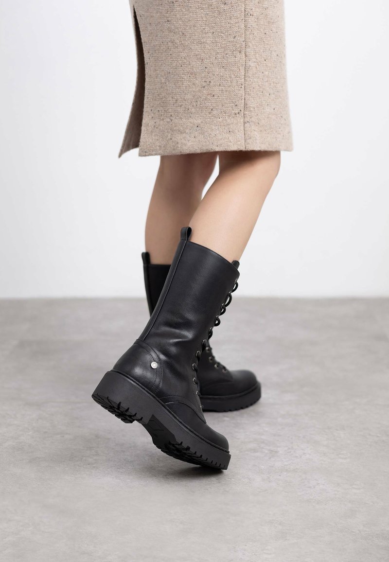 Botas de cuero negro con cordones y suela de goma gruesa, textura suave y tiradores laterales, combinadas con una falda de color marrón claro hasta la rodilla.
