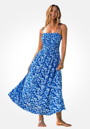 Lächelnde Frau trägt ein blaues, geblümtes Maxikleid mit dünnen Trägern und goldenen Sandalen mit Schnürung, hält den Saum des Kleides, während sie steht.