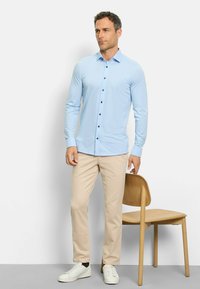 Chemise boutonnée bleu clair avec des boutons noirs, manches longues, associée à un pantalon beige et des baskets blanches. Chaise en bois en arrière-plan.