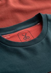 Donker teal en roestkleurige T-shirts gestapeld. Donker teal heeft een geribbelde halslijn; roestkleurig heeft een label met scharen en "KRAFT.COM" gedrukt.