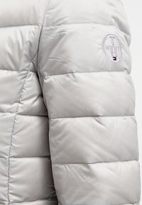 Jasnoszara, pikowana kurtka z rękawem, z naszywką w kształcie okręgu z haftowanymi literami "T N H" oraz małym emblematem flagi.