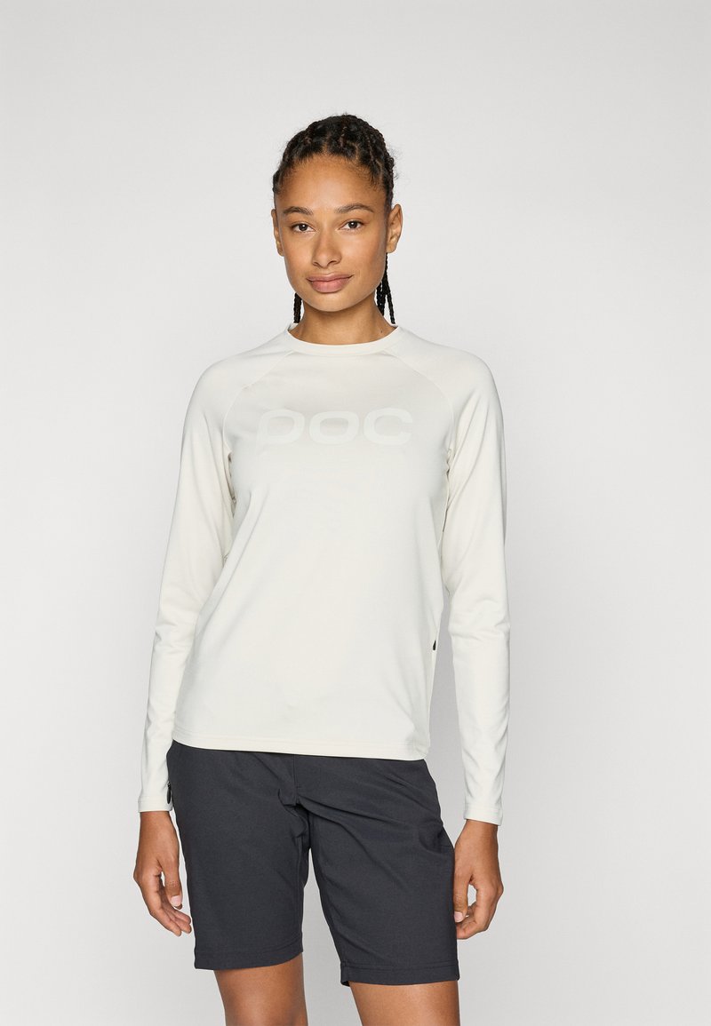 Chemise à manches longues de couleur beige clair en tissu lisse, arborant un grand logo subtil sur le devant. Assortie avec un short sombre.