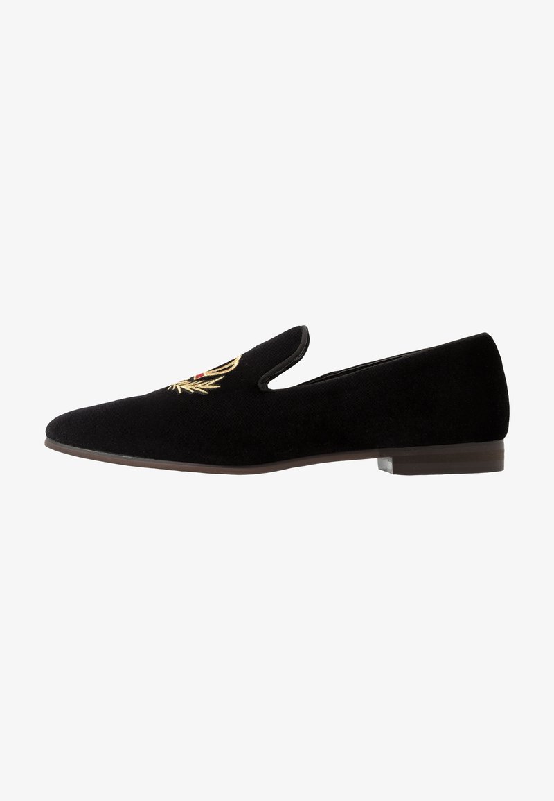 Chaussure slip-on en velours noir avec semelle plate et détail de couronne de laurier dorée brodée sur le dessus.