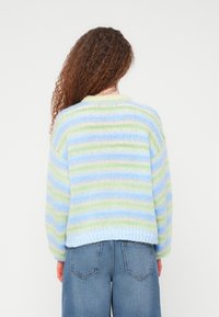 Γυναίκα με σγουρά μαλλιά που φοράει ένα πλεκτό πουλόβερ με ρίγες σε παστέλ μπλε, πράσινο και κρεμ χρώματα και μπλε τζιν παντελόνι, στέκεται μπροστά από ένα λευκό φόντο.