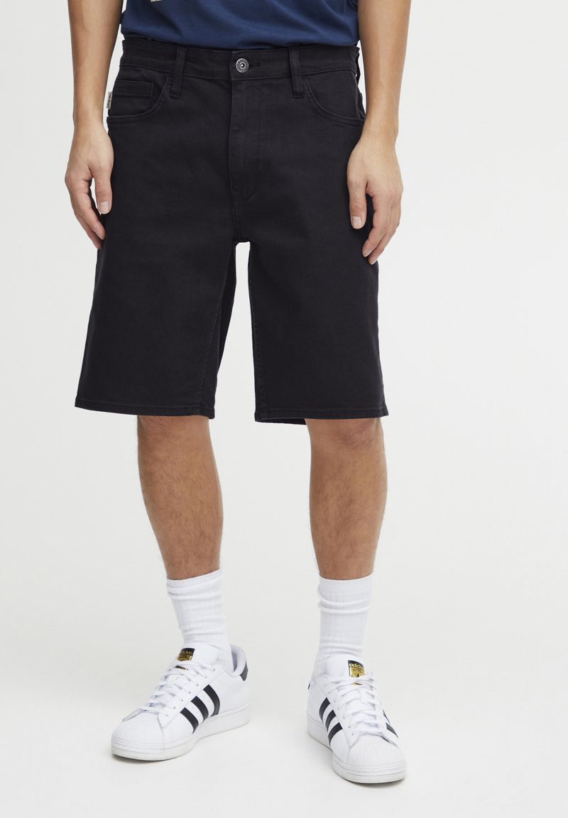Pantalones cortos de mezclilla negra con corte recto, cinco bolsillos y cierre de botón. Llevados con calcetas blancas y zapatillas negras a rayas.
